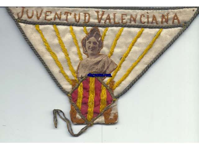 Juventud Valenciana.  Tringulo de tela bordado don se puede leer en la parte superior: Juventud Valenciana, seguido del busto de una mujer joven con el atuendo tradicional y acabado con un cuadradi representando las cuatro barras con una L a cada lado.